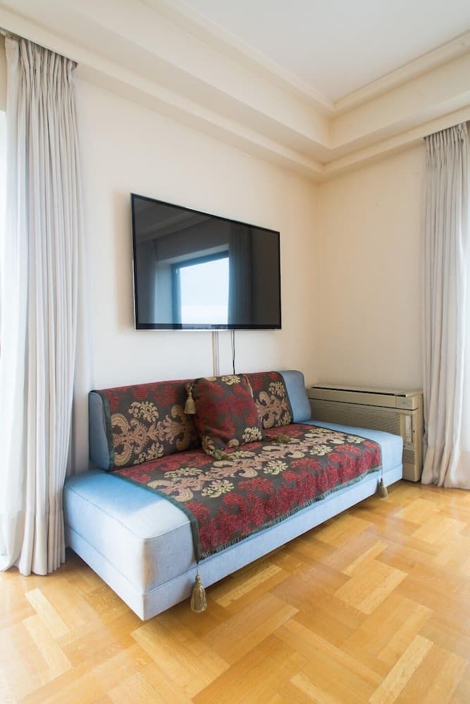 A cozy living room with a light blue sofa adorned with ornate red and gold textiles, a wall-mounted TV, wooden flooring, and soft curtains, ideal for travelers seeking a comfortable and stylish accommodation.