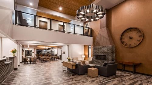 A cozy hotel lobby features warm wooden floors, modern furniture, a brick fireplace, and a large clock, creating an inviting atmosphere ideal for travelers seeking comfort and style.