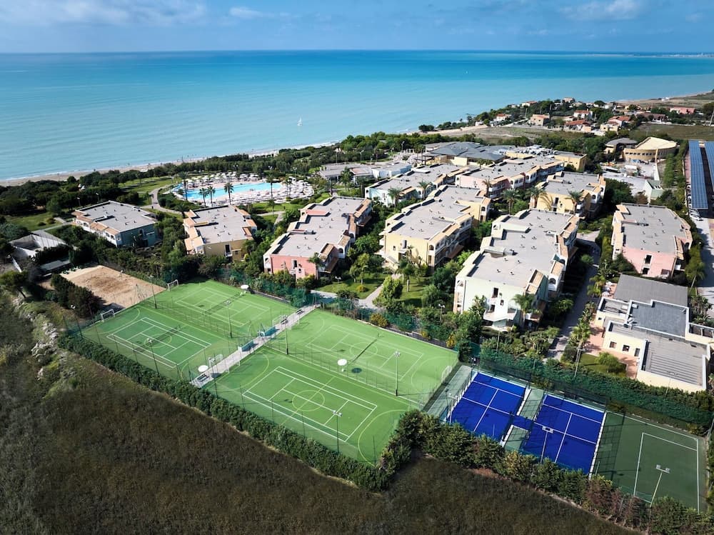 An aerial view of a vibrant coastal resort featuring tennis courts, swimming pools, and colorful buildings set against the serene backdrop of the ocean, ideal for family vacations and sports enthusiasts seeking relaxation and activity.