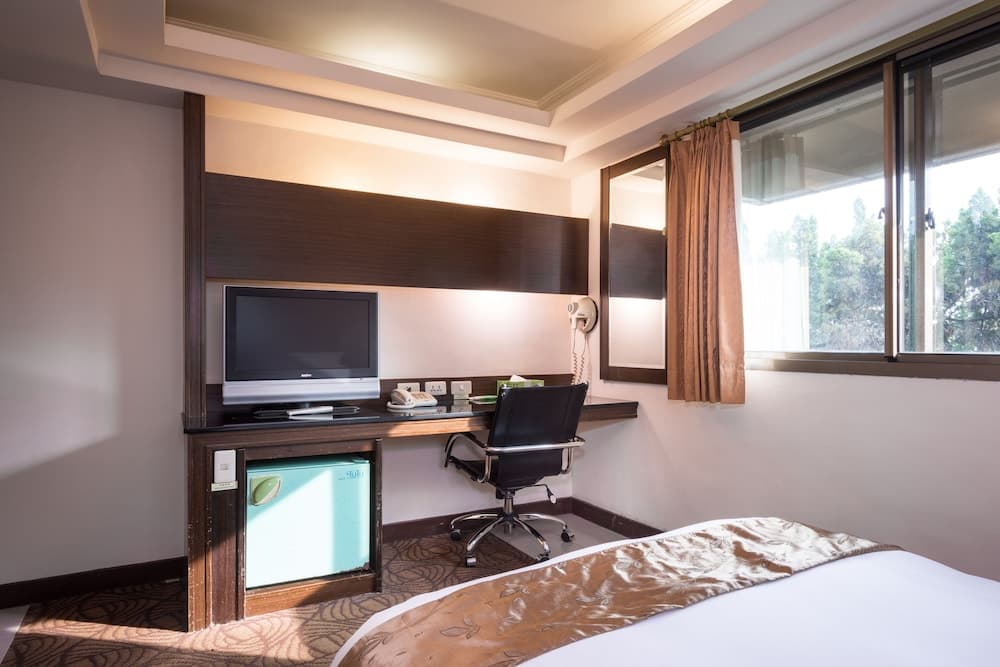 A cozy hotel room features a neatly made bed, a desk with a chair, a television, and a mini-fridge, set against a backdrop of soft lighting and a window view of greenery, ideal for travelers seeking comfort and convenience.
