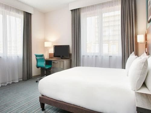 A cozy hotel room features a neatly made bed, a desk with a chair and TV, large windows with sheer curtains, and a warm ambiance, ideal for travelers seeking comfort and functionality during their stay.
