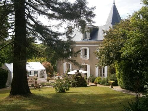 A serene countryside estate features a charming chateau surrounded by lush greenery, outdoor seating, and a white tent, ideal for a tranquil getaway or wedding venue.