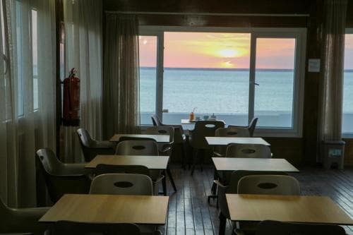 A serene dining room with wooden tables and chairs overlooks a breathtaking sunset over the ocean, ideal for travelers seeking a tranquil seaside retreat experience.