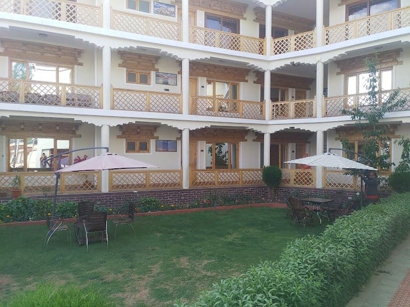 A serene courtyard at Hotel Ladakh Inn in Leh features lush greenery, cozy seating under umbrellas, and traditional wooden balconies, ideal for travelers seeking relaxation and cultural immersion.