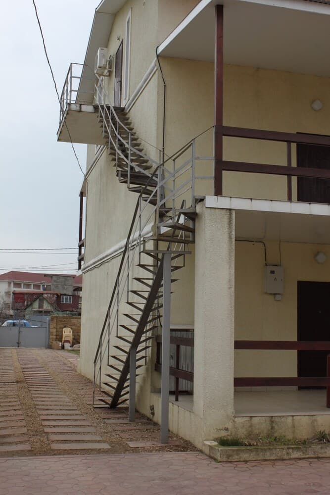 A quiet residential area features a beige building with an exterior spiral staircase, evoking a calm suburban mood ideal for travelers seeking peaceful accommodations.
