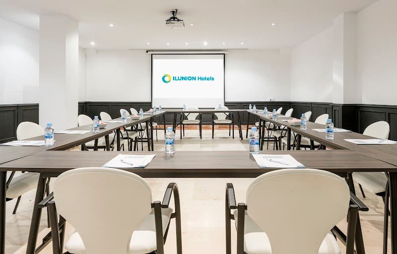 A modern conference room at Hotel ILUNION Bel Art in Barcelona features neatly arranged tables with bottled water, white chairs, and a projector screen displaying the hotel's logo, ideal for professional meetings and corporate events.