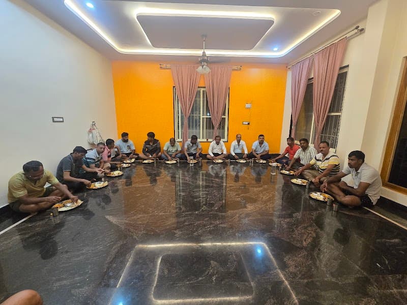 A group enjoys a communal meal in a warmly lit indoor space with polished floors and pink curtains, ideal for travelers seeking cultural experiences in Hampi, Karnataka.