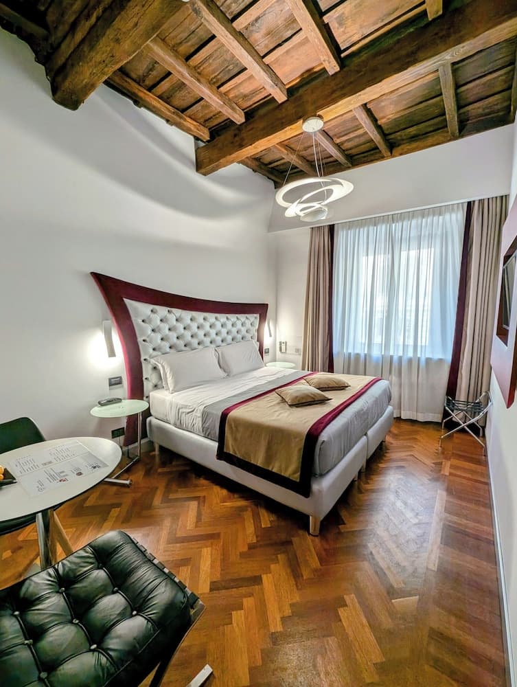 A cozy hotel room features a plush bed under an exposed wooden beam ceiling, complemented by elegant parquet flooring and modern lighting, ideal for travelers seeking a blend of rustic charm and contemporary comfort.