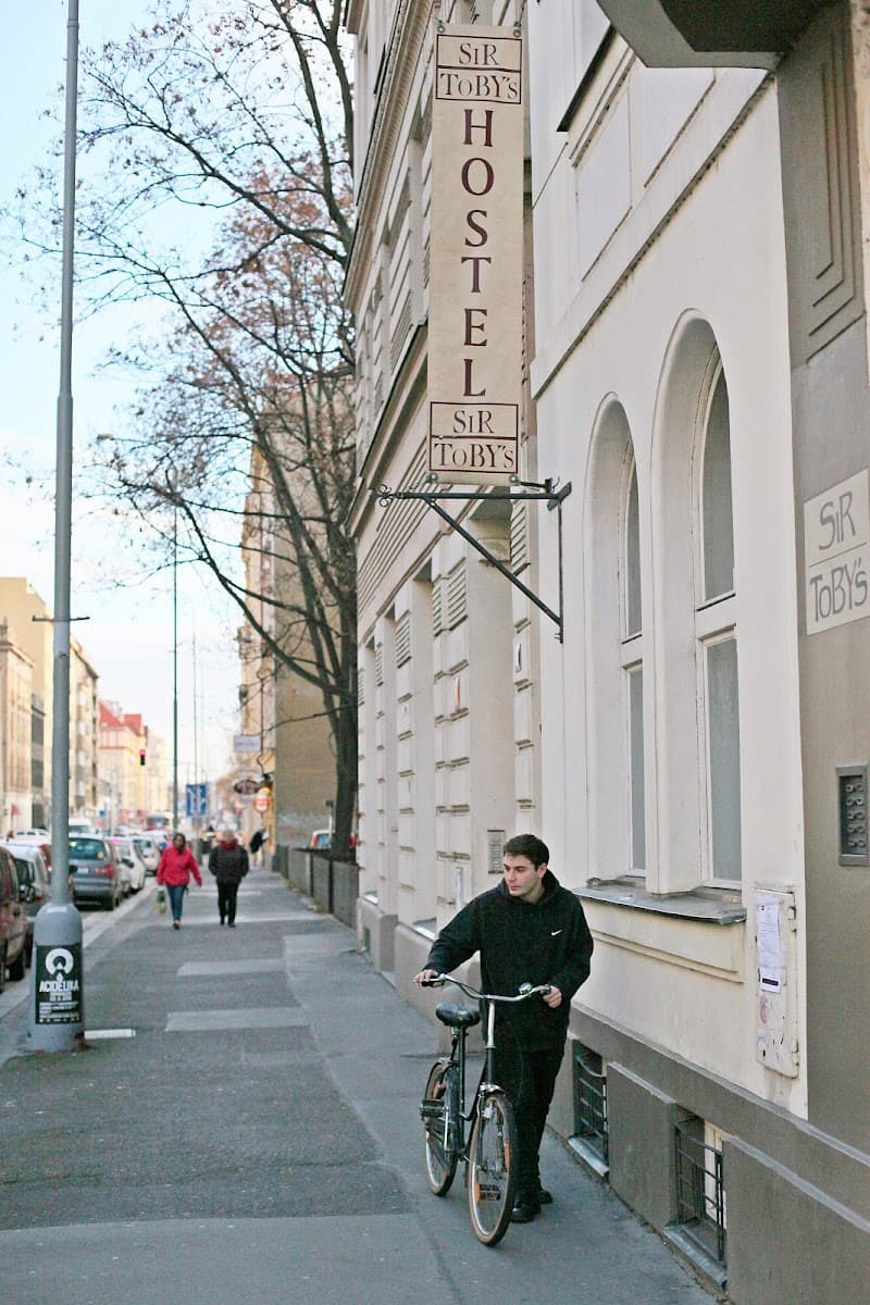 A cyclist pauses beside the charming Sir Toby's Hostel in Prague's Holešovice district, set against a backdrop of leafless trees and a quiet street, ideal for budget travelers exploring the city.
